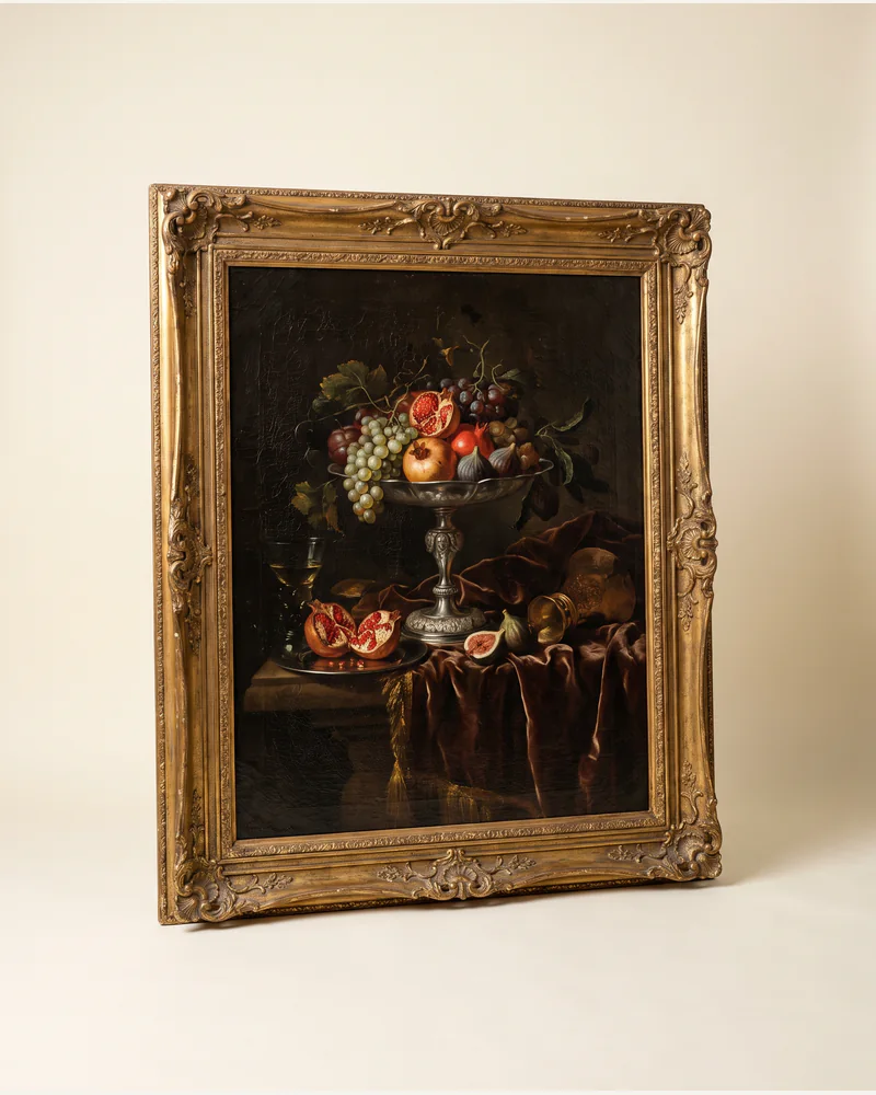 A dark Flemish still life of fruit, a silver tazza and drapery in a moulded gilt frame, photographed on a warm paper plate.